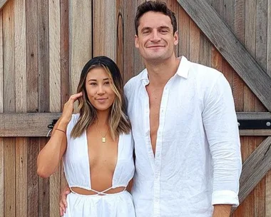 A couple in white outfits stands smiling in front of a wooden gate.