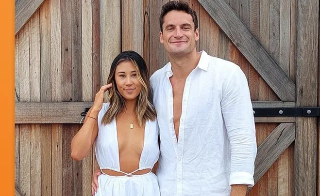 A couple in white outfits stands smiling in front of a wooden gate.