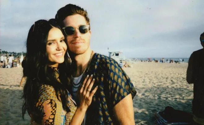 A couple smiling on a sunny beach, wearing patterned clothing and sunglasses.