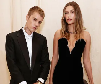 A couple stands side by side, he in a black suit and she in a black dress, against a neutral background.