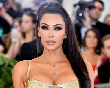 Woman with long dark hair in a gold dress, wearing cross necklaces, at an event with people and cameras in the background.