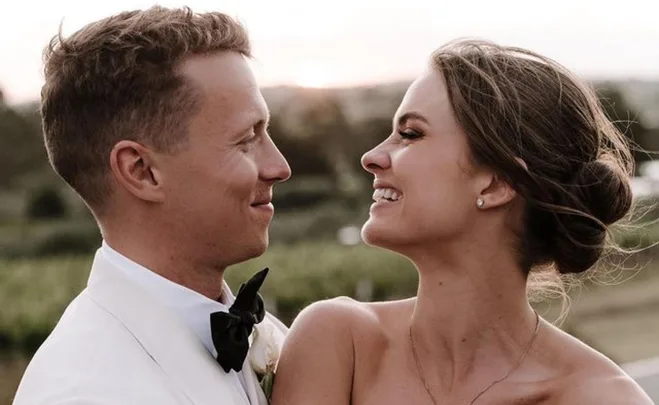 A couple smiling at each other outdoors during their wedding, with a sunset in the background.