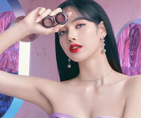 Lisa from BLACKPINK with bold makeup poses holding MAC eyeshadows, showcasing her collection.