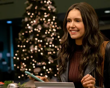 A woman smiling while holding a phone, with a blurred Christmas tree in the background.