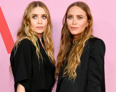 Two women with long, wavy blonde hair pose in black outfits against a pink background.