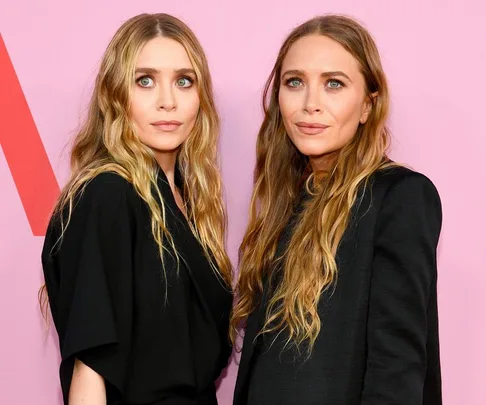 Two women with long, wavy blonde hair pose in black outfits against a pink background.