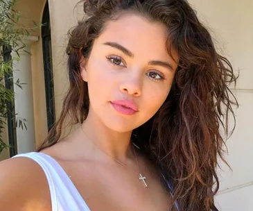 Young woman with wavy hair takes a selfie outside, wearing a white top and a cross necklace.