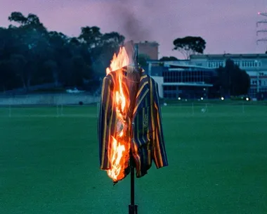 A striped blazer on fire stands in a field during dusk, with buildings and trees in the background.