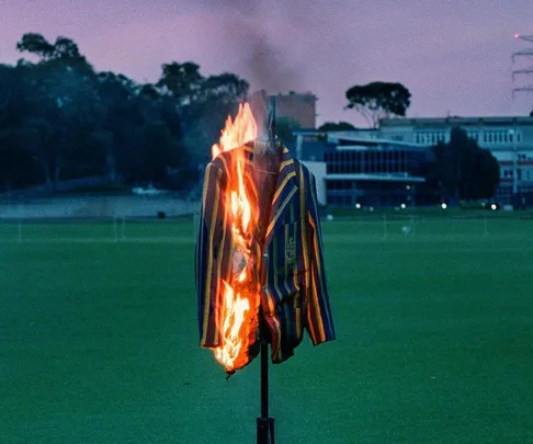 A striped blazer on fire stands in a field during dusk, with buildings and trees in the background.