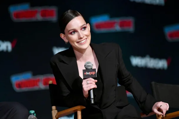 Person in a black blazer holding a microphone, smiling at a panel event with a "ReedPop" backdrop.