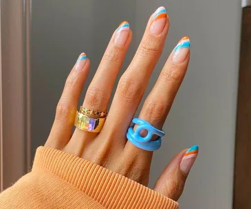 Colorful striped nail art on hand with gold and blue rings, orange sweater sleeve visible.