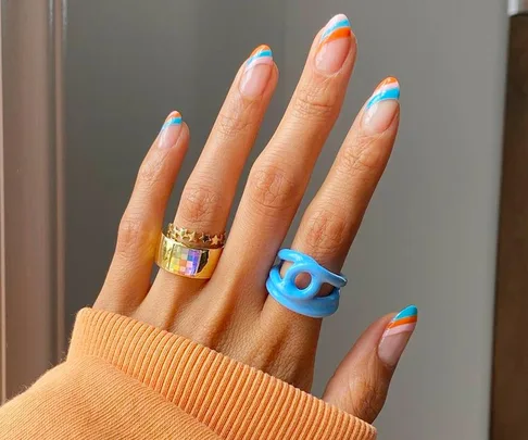 Colorful striped nail art on hand with gold and blue rings, orange sweater sleeve visible.