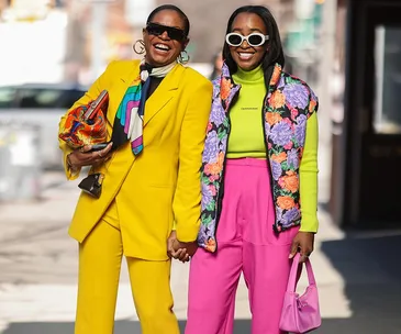 Two stylish women in colorful outfits smile and hold hands on a city street.