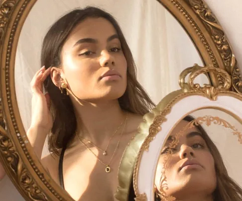 Woman wearing layered gold necklaces and earrings, looking in a vintage mirror, sunlight casting a warm glow.