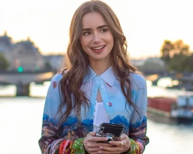 A woman with long brunette hair holds a camera, smiling by a riverside at sunset.