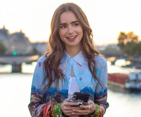 A woman with long brunette hair holds a camera, smiling by a riverside at sunset.