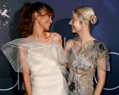 Two women in elegant dresses smiling at each other on a dark blue background.