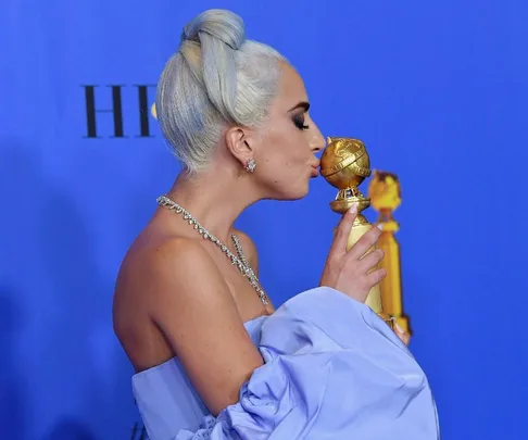 Woman with light hair kissing a Golden Globe award, wearing a light blue dress, against a blue background.