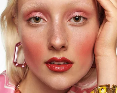 Close-up of a person with pink eye makeup, red lips, and geometric earrings, wearing a pink pattern outfit.