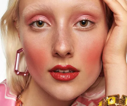 Close-up of a person with pink eye makeup, red lips, and geometric earrings, wearing a pink pattern outfit.