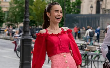 A woman in a red top and pink skirt smiles brightly while standing outside in a Parisian street setting.