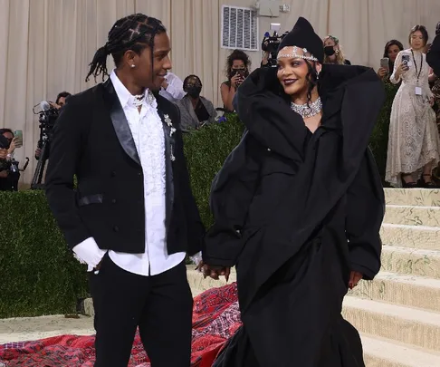 A couple in elegant black attire holding hands at a formal event, accompanied by photographers.