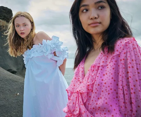 Two women in ruffled dresses stand on a rocky beach, one in white and one in pink, with a cloudy sky in the background.