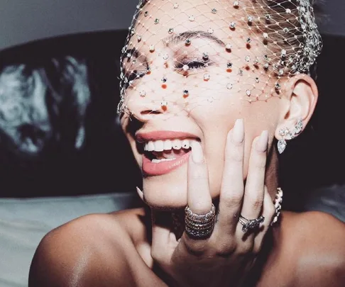 Smiling woman with jeweled veil and stylish tooth gems, adorned with rings and earrings.