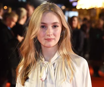 Young woman in a white blouse at a red carpet event with a blurred background.