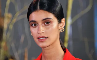 Woman with dark hair and gold earrings against a blurred background, wearing a bright red top.