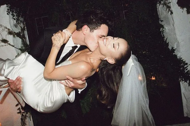 Bride in white dress being carried and kissed on cheek by groom in black suit under dim lights, smiling happily.