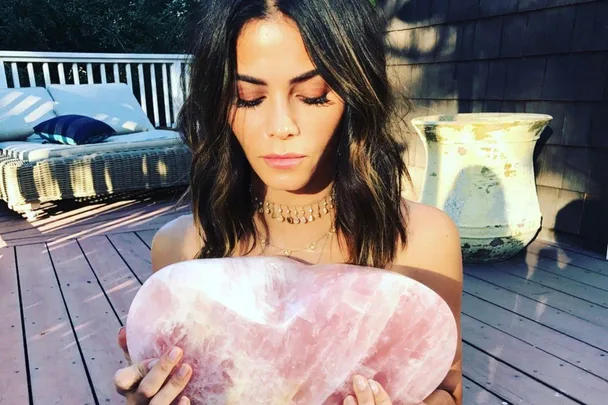 Woman holding a large, heart-shaped pink crystal on a sunny wooden deck.