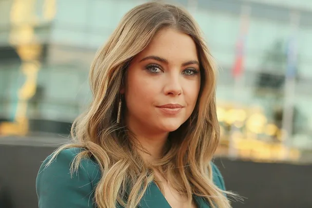 A person with long wavy hair and a serious expression, wearing a teal outfit, with a blurred background.