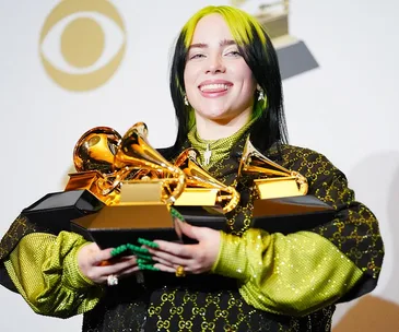 Billie Eilish smiling, holding multiple Grammy awards in a green and black Gucci outfit.