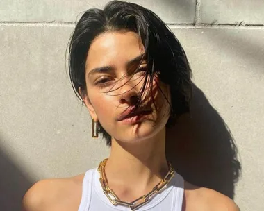 Woman with short bixie cut hair in sunlight, wearing a white top, gold chain necklace, and hoop earrings.