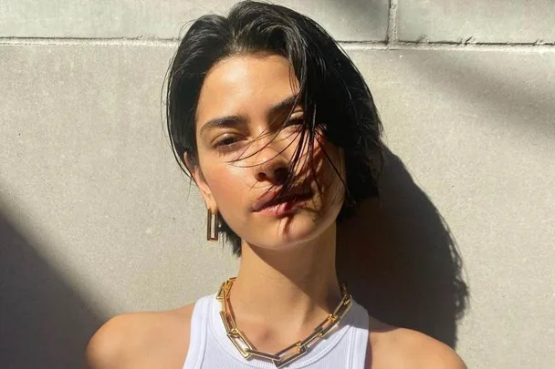 Woman with short bixie cut hair in sunlight, wearing a white top, gold chain necklace, and hoop earrings.