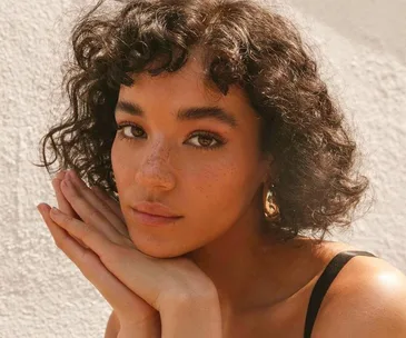 Person with curly hair and freckles, resting chin on hands, looking at the camera with a neutral expression.