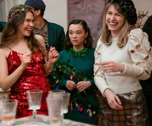 Three women in festive outfits at a holiday party, smiling and laughing near a table with empty glasses.