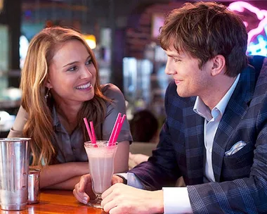 A couple smiling at each other over a shared milkshake with pink straws in a cozy restaurant setting.