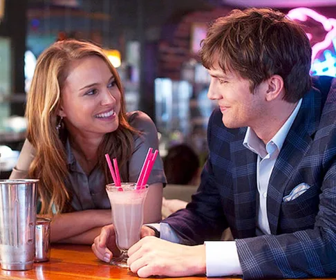 A couple smiling at each other over a shared milkshake with pink straws in a cozy restaurant setting.