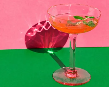 A cocktail in a glass with mint garnish, set against a pink and green background.