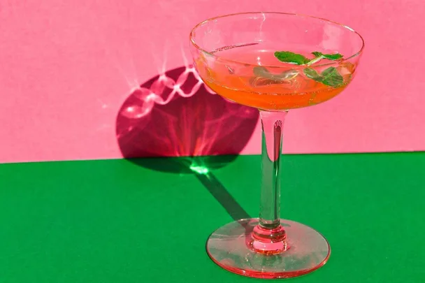 A cocktail in a glass with mint garnish, set against a pink and green background.