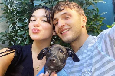 A couple poses for a selfie with a small black dog, greenery in the background.