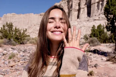 Woman smiling and showing off her engagement ring in a desert landscape.