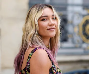 A woman with blonde and pink-tinted hair, wearing a colorful floral dress, looks to the side outdoors.