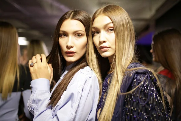 Two women with long hair stand close together, wearing blue outfits in a dimly lit room.