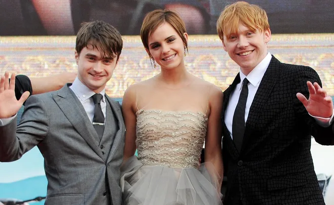 Three cast members from Harry Potter stand together, smiling and waving at an event.