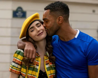 A man kissing a smiling woman on the cheek while embracing her; she wears a yellow beret and plaid outfit.