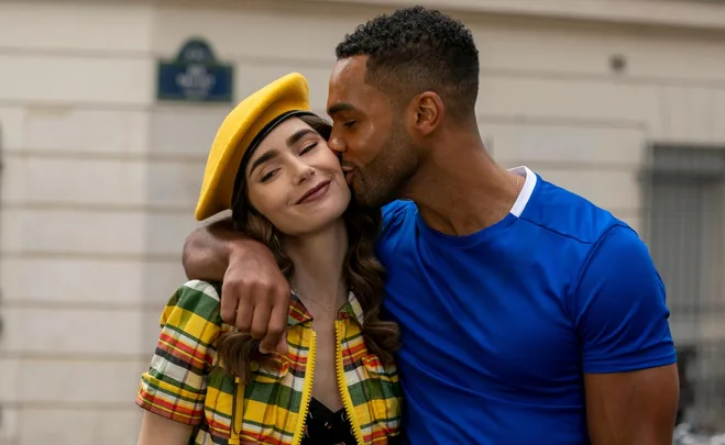 A man kissing a smiling woman on the cheek while embracing her; she wears a yellow beret and plaid outfit.