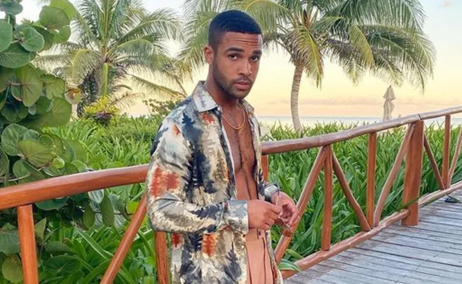 Man in patterned shirt stands by wooden railing with tropical plants and palm trees in the background.